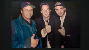 "Three men posing together, all smiling and giving thumbs up; two wear baseball caps and casual jackets, while one in the middle is in a dark blazer. The background is dark, suggesting an indoor or nighttime setting."